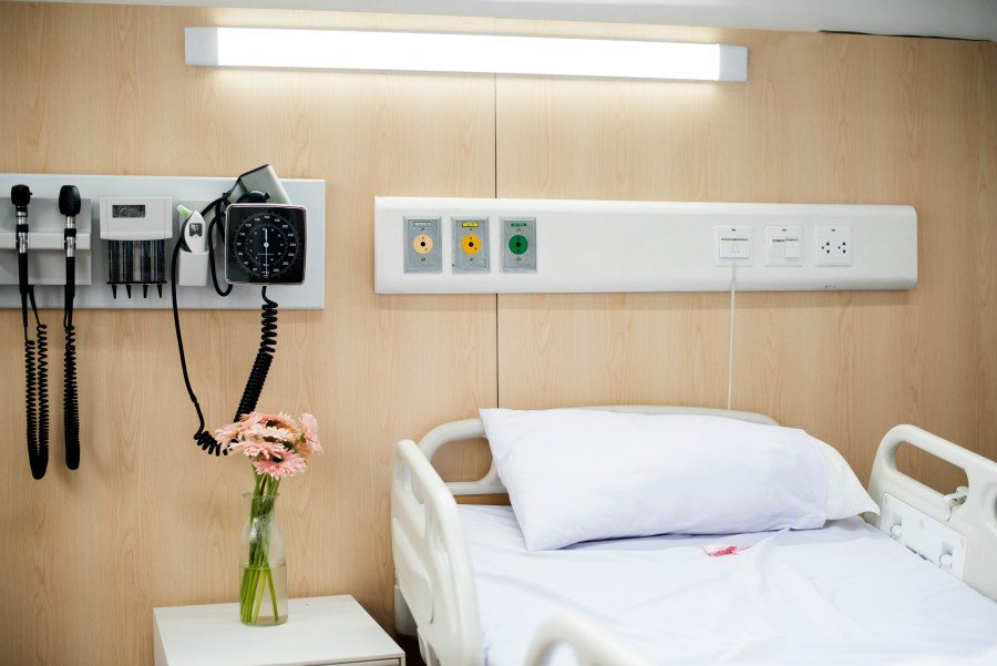 Hospital patient bed with white linens, bedside medical equipment, wall-mounted outlets, and a vase of pink flowers.