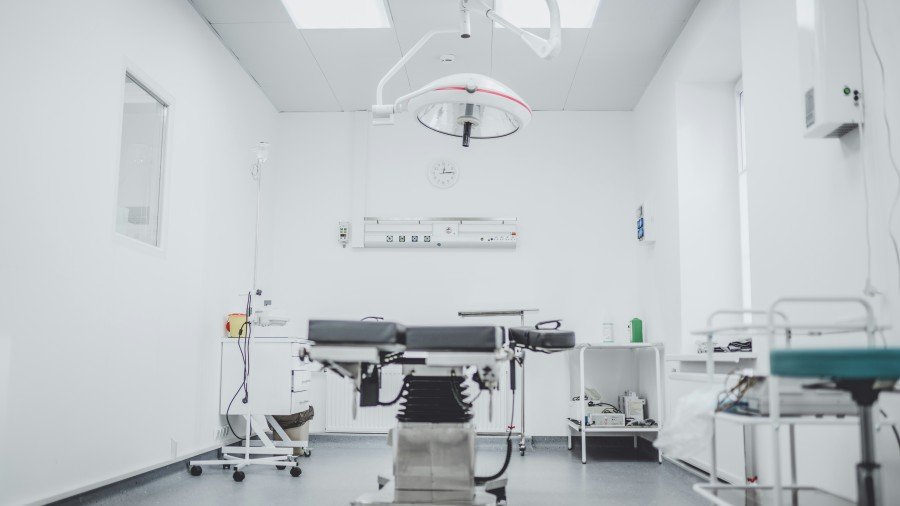 Bright, modern operating room with a surgical table, overhead surgical light, medical equipment, and storage carts.