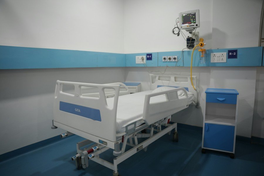 Hospital room with an adjustable patient bed, bedside cabinet, wall-mounted medical monitor, oxygen equipment, and blue-and-white furnishings.
